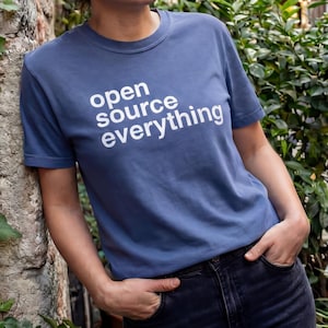 May include: A blue t-shirt with the white text "open source everything". The person is wearing dark blue jeans. The person is standing near a tree trunk covered in ivy. The background shows a street scene.