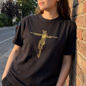 Könnte beinhalten: Schwarzes T-Shirt mit einem goldenen Leiterplattendesign in Form eines Kreuzes. Das kurzärmelige Oberteil hat einen Rundhalsausschnitt. Die Person lehnt an einer Backsteinmauer.