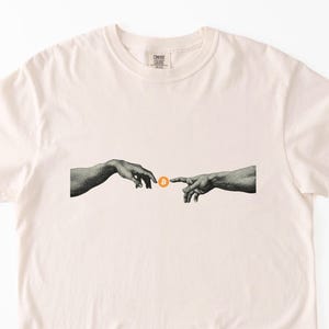 May include: Cream-colored t-shirt featuring a graphic of two hands reaching towards each other, with a Bitcoin symbol between them. The design is reminiscent of the Creation of Adam. The shirt has a classic crew neck and short sleeves.