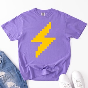 Puede incluir: Una camiseta morada con un diseño de rayo amarillo pixelado. La imagen también muestra unos vaqueros azules y zapatillas blancas, sugiriendo un estilo relajado e informal. La camiseta es de un material suave.