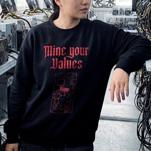 Könnte beinhalten: Schwarzes Sweatshirt mit rotem Aufdruck "Mine your Values" in gotischer Schrift. Darunter befindet sich eine rote Grafik einer Platine mit dem Wort "Bitaxe".