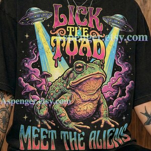 May include: Black graphite t-shirt featuring a colorful graphic of a frog with the text "Lick the Toad, Meet the Aliens." The design includes UFOs, mushrooms, and vibrant colors. The shirt is from Comfort Colors.