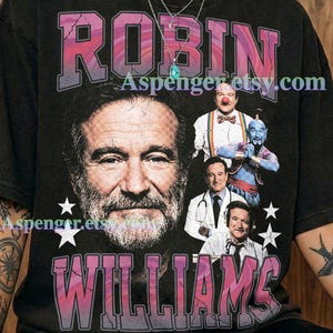 May include: A graphite-colored t-shirt featuring a large portrait of Robin Williams, the text "ROBIN WILLIAMS" in a cracked pink and purple font, and smaller images of the actor in various roles, including the Genie and a doctor, with white stars.