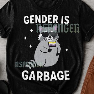 Può includere: T-shirt unisex nera con la scritta "GENDER IS GARBAGE". La maglietta presenta un procione dei cartoni animati con occhiali da sole e una bandiera non binaria. La maglietta ha anche la scritta "UNISEX TSHIRT" su una piccola etichetta bianca.