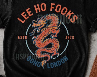 Może przedstawiać: Czarny, uniseksowy t-shirt z postarzaną grafiką przedstawiającą pomarańczowego smoka. Tekst "LEE HO FOOKS" znajduje się nad smokiem, z "ESTD .1978" i "SOHO LONDON" poniżej, w niebieskim okręgu.