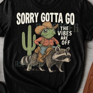 May include: Black unisex t-shirt with a humorous graphic. The design features a frog in a cowboy hat riding a raccoon, with a cactus and the text "SORRY GOTTA GO THE VIBES ARE OFF".