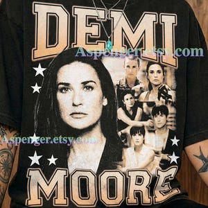 May include: A graphite-colored t-shirt featuring a large graphic print of Demi Moore. The design includes the name DEMI MOORE in a bold, collegiate-style font, multiple black and white photographic portraits of the actor, and white star icons.