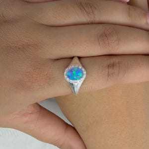 May include: A silver ring with an oval, blue opal stone, surrounded by a halo of small, clear stones. The ring is set on a hand with pink nail polish. The ring's design is elegant and eye-catching, suitable for everyday wear or special occasions.