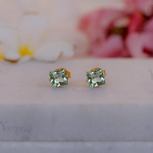 May include: Two square-cut, pale green gemstone stud earrings with gold settings. The earrings are presented on a white surface, with blurred flowers in the background. The earrings are a delicate piece of jewellery.