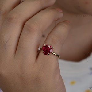 Op de afbeelding: Een gouden ring met een gefacetteerde, hartvormige rode edelsteen die door vier pootjes wordt vastgehouden. De ringband is van glad, gepolijst goud. De tekst YOROMA is zichtbaar en verschijnt meerdere keren als watermerk in de afbeelding en hoek.