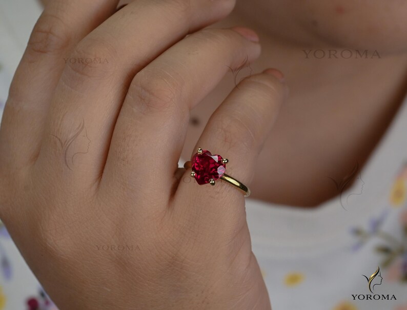 Puede incluir: Un anillo de oro con una piedra preciosa roja en forma de coraz&oacute;n facetado, sujeta por cuatro garras. El aro es de oro liso y pulido. El texto YOROMA es visible, apareciendo varias veces como marca de agua en la imagen y en la esquina.