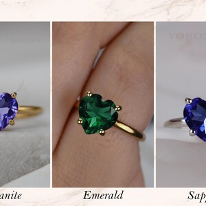 Puede incluir: Tres anillos con piedras preciosas en forma de coraz&oacute;n etiquetados como YOROMA. El primero es una tanzanita morada en banda dorada, el segundo una esmeralda verde en banda dorada y el tercero un zafiro azul en banda plateada, cada uno con cuatro garras.