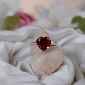 Puede incluir: Un anillo de plata con una piedra preciosa roja tallada en forma de coraz&oacute;n y sujeta por cuatro garras. El anillo est&aacute; colocado sobre una concha espiral de color claro, situada entre pliegues de tela de sat&eacute;n blanca. El texto YOROMA es visible.