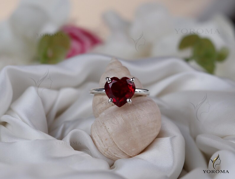 Puede incluir: Un anillo de plata con una piedra preciosa roja tallada en forma de coraz&oacute;n y sujeta por cuatro garras. El anillo est&aacute; colocado sobre una concha espiral de color claro, situada entre pliegues de tela de sat&eacute;n blanca. El texto YOROMA es visible.