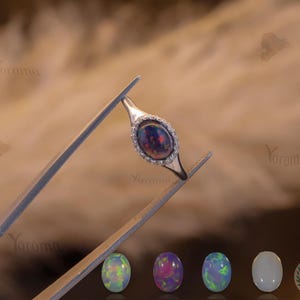 Op de afbeelding: Een zilveren ring met een grote, ovale, zwarte opaal edelsteen, omringd door kleine, heldere stenen. De ring wordt vastgehouden door een pincet. Daaronder bevinden zich vier ovale edelstenen in verschillende kleuren, waaronder groen, paars en wit.