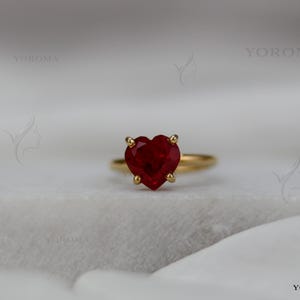 Puede incluir: Un anillo de oro con una piedra preciosa roja tallada en forma de coraz&oacute;n y sujeta por cuatro garras. La palabra YOROMA es visible en la imagen. El anillo tiene una banda de oro fina y lisa que rodea el dedo, mostrando un dise&ntilde;o cl&aacute;sico.