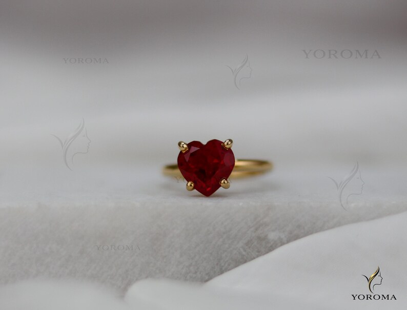 Puede incluir: Un anillo de oro con una piedra preciosa roja tallada en forma de coraz&oacute;n y sujeta por cuatro garras. La palabra YOROMA es visible en la imagen. El anillo tiene una banda de oro fina y lisa que rodea el dedo, mostrando un dise&ntilde;o cl&aacute;sico.