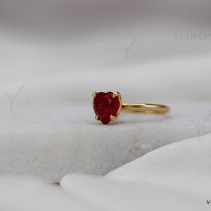 Puede incluir: Un anillo de oro con una piedra preciosa roja facetada en forma de coraz&oacute;n, montada en un engaste de cuatro garras. El aro es de oro liso y pulido. El texto YOROMA es visible en las esquinas superior e inferior derecha, junto a un logo.
