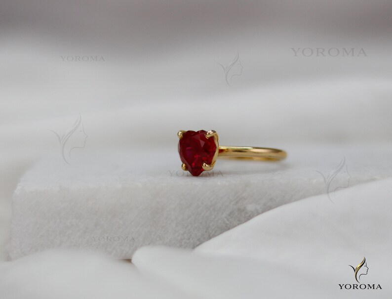 Puede incluir: Un anillo de oro con una piedra preciosa roja facetada en forma de coraz&oacute;n, montada en un engaste de cuatro garras. El aro es de oro liso y pulido. El texto YOROMA es visible en las esquinas superior e inferior derecha, junto a un logo.