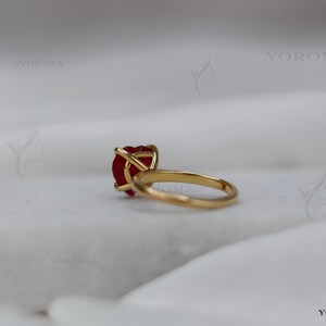 Puede incluir: Un anillo de oro con una piedra preciosa roja en forma de coraz&oacute;n sujeta por un engaste de cuatro garras. El aro es liso y circular, con un acabado met&aacute;lico pulido. La palabra YOROMA es visible como marca de agua en varias ubicaciones.