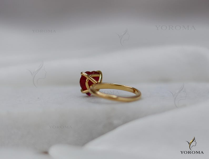Puede incluir: Un anillo de oro con una piedra preciosa roja en forma de coraz&oacute;n sujeta por un engaste de cuatro garras. El aro es liso y circular, con un acabado met&aacute;lico pulido. La palabra YOROMA es visible como marca de agua en varias ubicaciones.