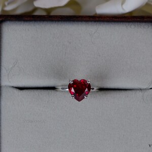 Puede incluir: Un anillo de plata con una piedra preciosa roja tallada en forma de coraz&oacute;n y sujeta por cuatro garras. El anillo est&aacute; centrado en una caja de presentaci&oacute;n cuadrada de bordes oscuros con un interior gris claro texturizado. El nombre YOROMA es visible.