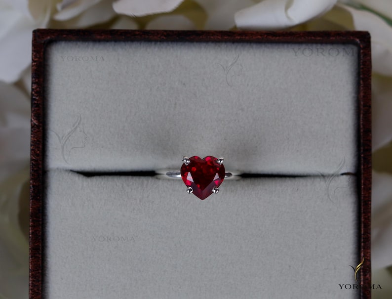 Puede incluir: Un anillo de plata con una piedra preciosa roja tallada en forma de coraz&oacute;n y sujeta por cuatro garras. El anillo est&aacute; centrado en una caja de presentaci&oacute;n cuadrada de bordes oscuros con un interior gris claro texturizado. El nombre YOROMA es visible.