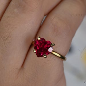 Puede incluir: Un anillo de oro con una piedra preciosa roja tallada en forma de coraz&oacute;n y sujeta por cuatro garras. El aro es de oro liso y pulido. El texto YOROMA es visible en la imagen, apareciendo varias veces como una marca de agua.