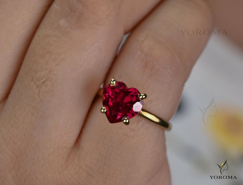 Puede incluir: Un anillo de oro con una piedra preciosa roja tallada en forma de coraz&oacute;n y sujeta por cuatro garras. El aro es de oro liso y pulido. El texto YOROMA es visible en la imagen, apareciendo varias veces como una marca de agua.