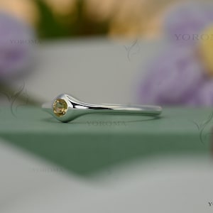 May include: A silver ring featuring a round, pale-yellow gemstone. The ring has a smooth, polished band and a bezel setting. The ring is displayed on a green surface with blurred flowers in the background. The ring is from the brand Yoroma.