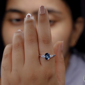 Può includere: Un anello d'argento con una pietra preziosa ovale blu scuro e due pietre preziose più piccole e trasparenti. L'anello è su un dito, con uno sfondo sfocato.