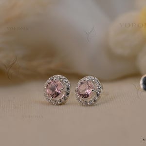 May include: A pair of stud earrings featuring a round, light pink center stone surrounded by a halo of small, clear white stones set in a silver-toned metal. The earrings include silver-toned metal backings and the text YOROMA is visible.