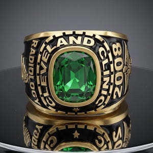 Può includere: Un anello color oro con una grande pietra rettangolare sfaccettata verde smeraldo al centro. La lunetta in smalto nero è incisa con il testo CLEVELAND CLINIC RADIOLOGY 2008, accentuata da piccole stelle e emblemi di aquila sui lati.