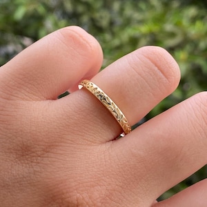 Könnte beinhalten: Ein goldener Ring mit einem komplizierten Blattmuster. Der Ring ist dünn und scheint aus poliertem Metall zu bestehen. Der Ring befindet sich an einem Finger, mit einem verschwommenen grünen Hintergrund.