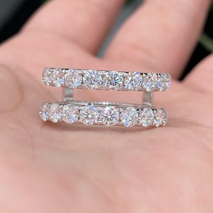 May include: Two silver rings with a row of clear, sparkling gemstones. The rings are stacked, with one above the other. The gemstones are round and set closely together, creating a continuous band of light.
