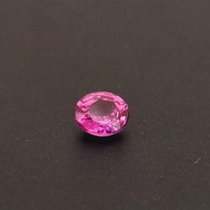 Peut inclure: Une pierre précieuse rose taillée en forme ovale. La gemme est d'une teinte rose vif, avec de multiples facettes qui réfléchissent la lumière. La pierre est posée sur un fond sombre, ce qui met en valeur sa couleur et sa brillance.