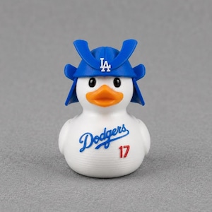 May include: A white rubber duck wearing a blue samurai-style helmet with a white LA logo on the front. The duck features an orange beak, black eyes, and the blue script text Dodgers with the red number 17 printed on its chest area.