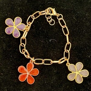 May include: Gold-toned chain bracelet with three flower charms. The charms are in shades of purple, orange, and gray, with gold-colored outlines. The bracelet has a lobster clasp closure.