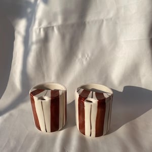 May include: Two ceramic containers with a white base and vertical maroon stripes. Each container has a small opening at the top. The containers are cylindrical and sit on a white surface. The containers are handmade.