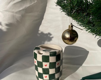 Puede incluir: Una taza de cerámica con un patrón de cuadros verdes y blancos y pequeños acentos de corazón rojo. La taza cilíndrica tiene una parte superior redondeada y está sobre un fondo blanco. Un árbol de Navidad y una bola dorada están en el fondo.