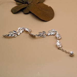 May include: A silver-colored bracelet featuring a delicate leaf design. The bracelet has multiple leaf-shaped charms connected by a chain, with a small ball at the end. The jewelry is displayed on a neutral background.