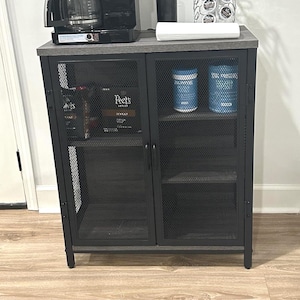 May include: A black and gray coffee station with a Cuisinart coffee maker, a black canister, and a K-cup holder. The cabinet has a metal mesh door and contains coffee and protein powder.