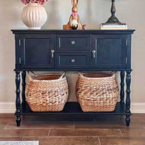 May include: A black cabinet with two doors and two drawers, topped with a vase of pink flowers, a bunny figurine holding a golden egg, and a stained glass lamp. Two woven baskets sit below.