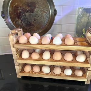 May include: A three-tiered wooden egg holder filled with various colored eggs. The eggs range in color from light tan to white. The holder is made of light-colored wood and has a rustic appearance. The background includes kitchen items.