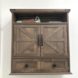 May include: A rustic-style wall-mounted cabinet with a distressed wood finish. It features two doors with a diagonal cross design, two drawers, and black hardware. The cabinet has an open top shelf.