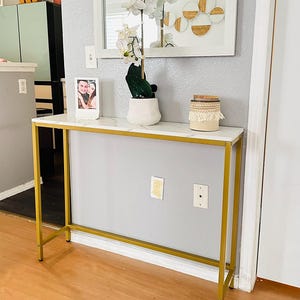 May include: A gold metal console table with a white marble top. The table has a rectangular shape and is supported by four slender legs. A framed photo, a white potted plant with an orchid, and a decorative jar are on the table.