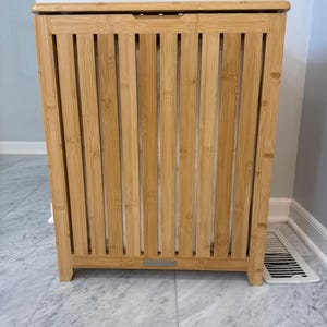 May include: A natural bamboo laundry hamper with a slatted front and a hinged lid. The hamper is rectangular with a light brown finish and a small metal label on the front. It stands on four short legs.