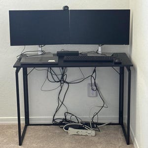 May include: A black computer desk with two black monitors, a laptop, keyboard, and mouse. The desk has a black metal frame and is against a white wall. Cables are visible under the desk, and a power strip rests on the floor.