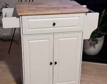 May include: A white kitchen island with a natural wood countertop. It features a drawer, two cabinet doors, and a pull-out shelf. The island is on wheels for easy mobility.