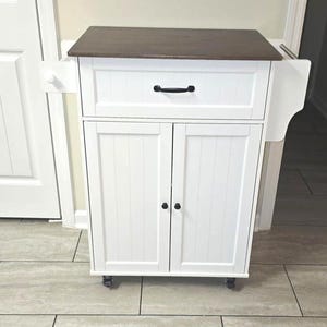 May include: A white kitchen cart with a dark brown countertop. The cart has a drawer with a black handle and two cabinet doors. The cart is on wheels and has a towel rack on the side.